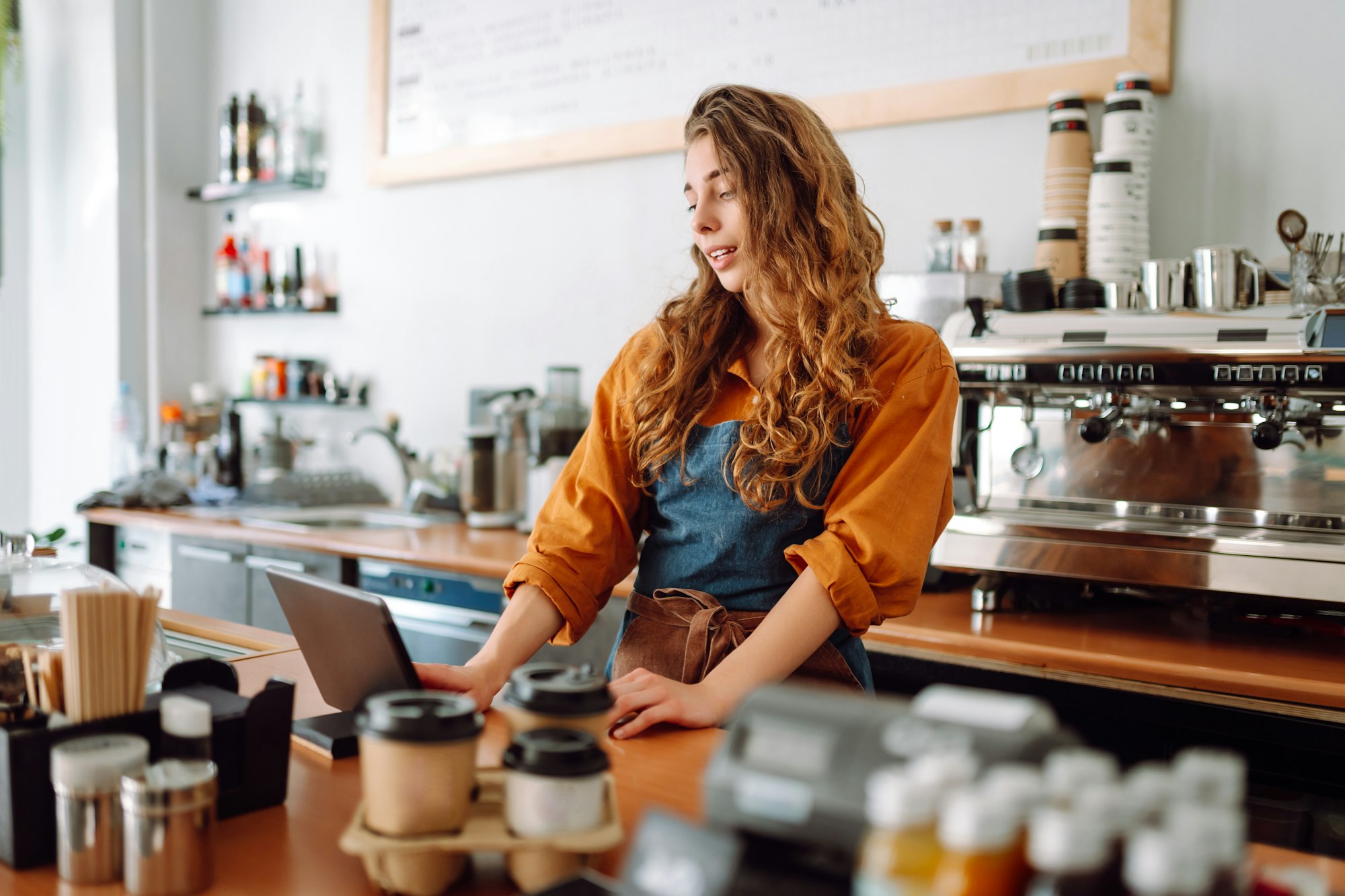 Young woman in apron with digital tablet at bar counter of coffee shop takes order. Small business.
