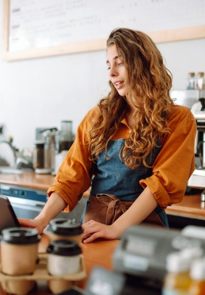 Young woman in apron with digital tablet at bar counter of coffee shop takes order. Small business.